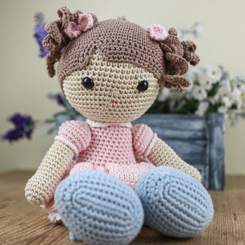 Close da boneca Mari de crochê, com cabelo de cachos e vestido rosa, diante de um arranjo de flores