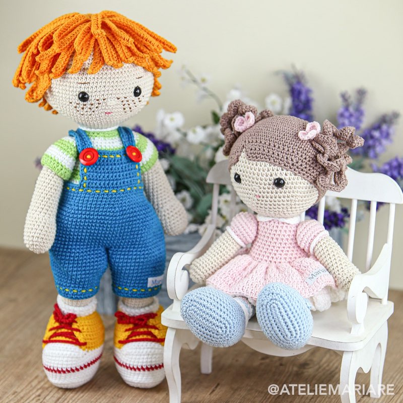 Boneco de crochê com cabelo cacheado laranja, em pé, ao lado de uma boneca com cabelo castanho e vestido rosa, sentada em um banquinho branco entre flores