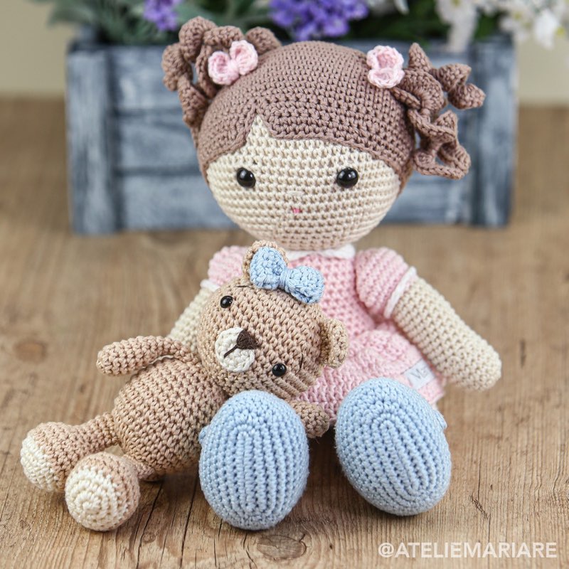Boneca de crochê com cabelo castanho cacheado e vestido rosa, segurando um ursinho de amigurumi com laço azul