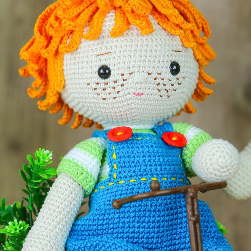 Boneco de crochê com cabelo cacheado laranja e sardinhas, vestindo jardineira azul, ao lado do guidão de uma bicicleta em miniatura