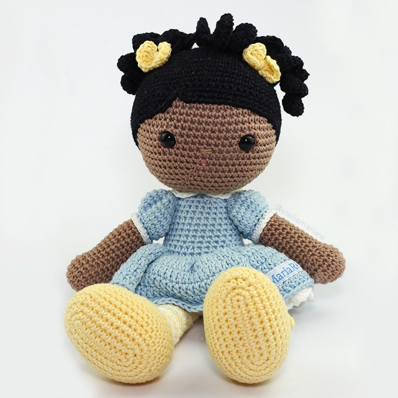 Boneca de crochê de pele morena com cabelo cacheado preso e vestido azul claro