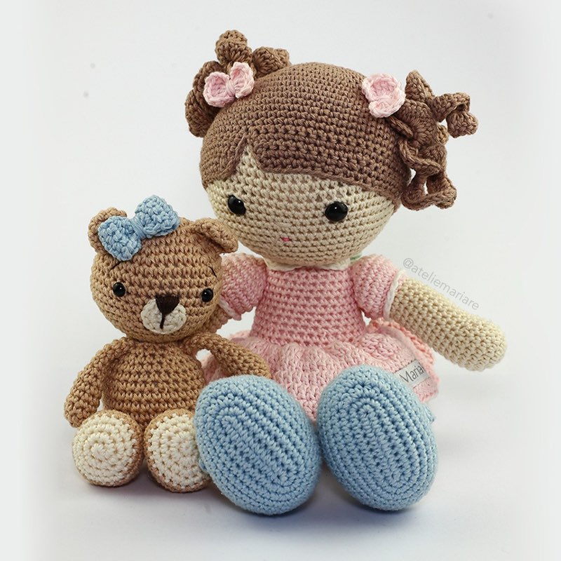 Boneca de crochê com cabelo castanho e vestido rosa, sentada ao lado de um ursinho de amigurumi com laço azul