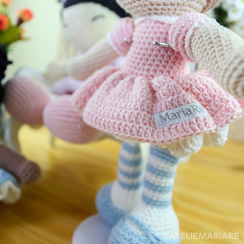 Boneca de crochê com vestido rosa e pernas listradas azul e branco, em pé com etiqueta artesanal