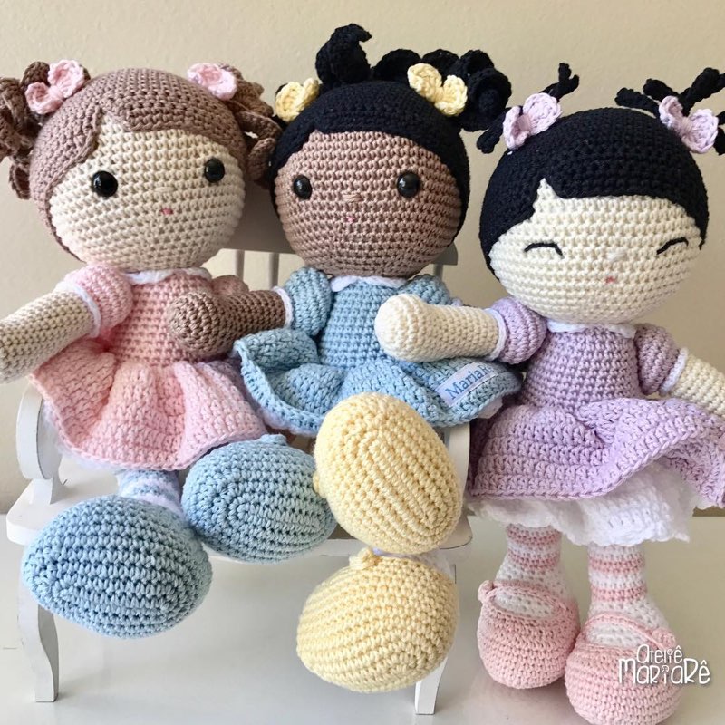 Três bonecas de crochê com vestidos rosa, azul e lilás, abraçadas em um banquinho