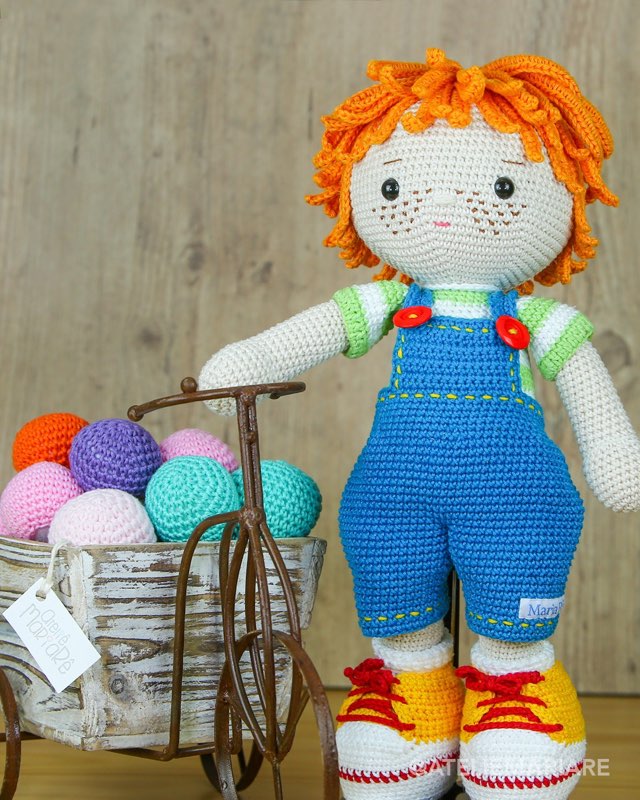 Boneco de crochê com cabelo laranja cacheado e jardineira azul, em pé ao lado de uma cesta de novelos coloridos