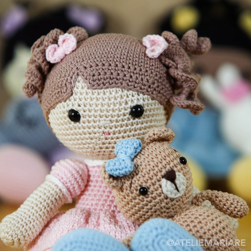 Boneca de crochê com laço azul sentada ao lado de um ursinho de amigurumi, em cima da cama
