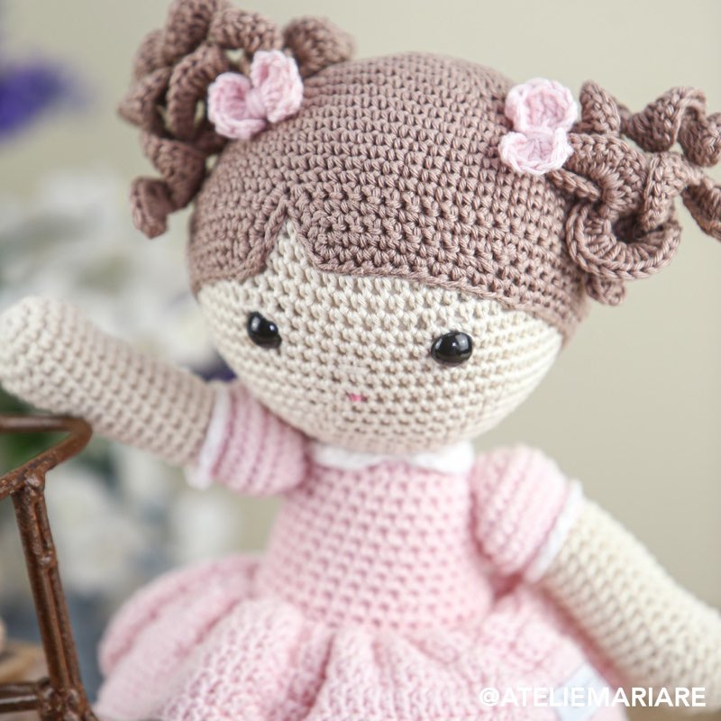 Boneca de crochê com cabelo cacheado e vestido rosa, em pé em um jardim de madeira