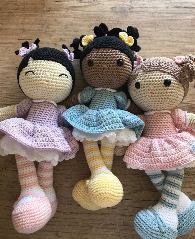 Três bonecas de crochê com vestidos de bailarina rosa, azul e amarelo, em tons de pele diferentes