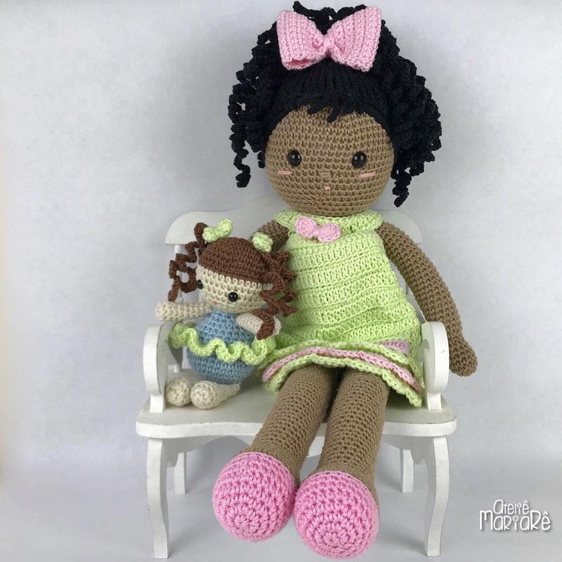 Boneca de crochê de pele morena com vestido verde, sentada em um banquinho branco segurando uma bonequinha bebê