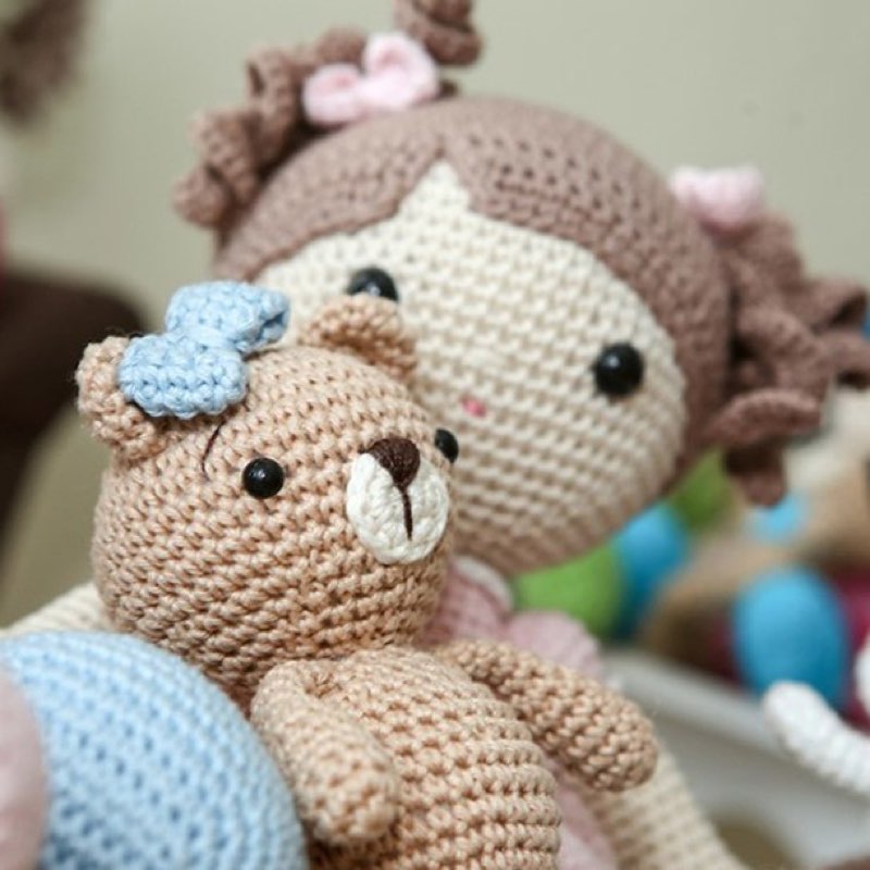 Boneca de crochê com laço azul abraçada a um ursinho de amigurumi, deitados na cama
