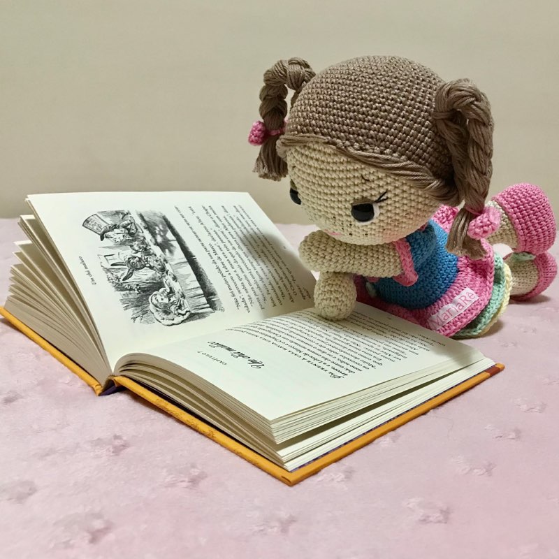 Boneca de crochê sentada lendo um livro aberto, com as pernas apoiadas nas páginas