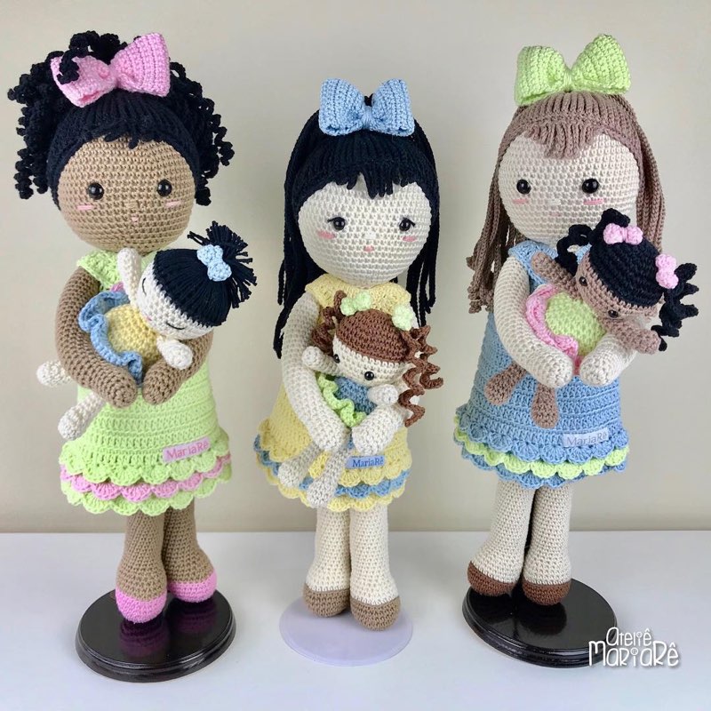 Três bonecas de crochê de tons de pele diferentes, cada uma segurando uma bonequinha ou bichinho pequeno