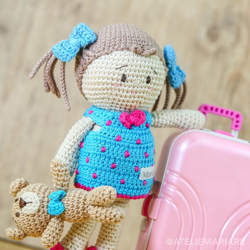 Boneca de crochê com roupinha azul e mochila rosa, ao lado de um ursinho pequeno de amigurumi