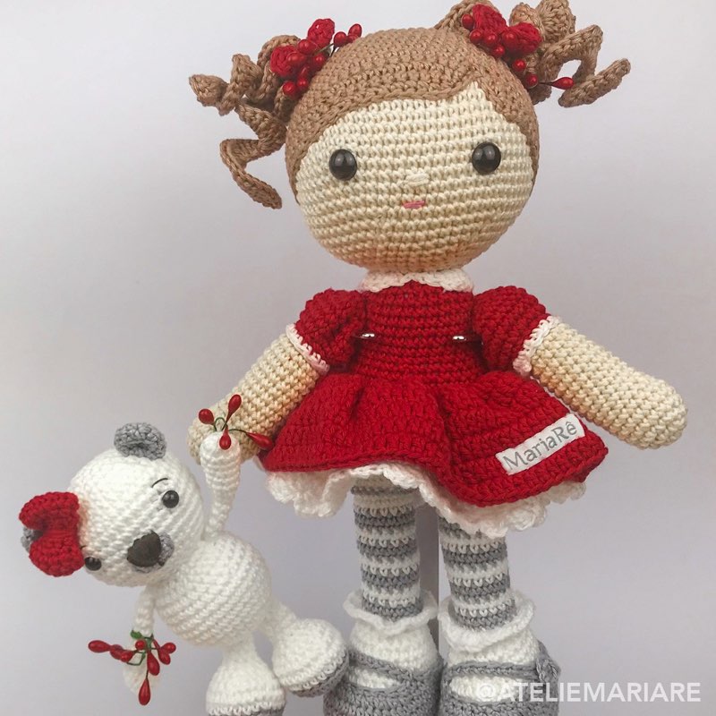 Boneca de crochê com vestido vermelho de Natal ao lado de um cordeiro branco de amigurumi com laço vermelho e bagas natalinas