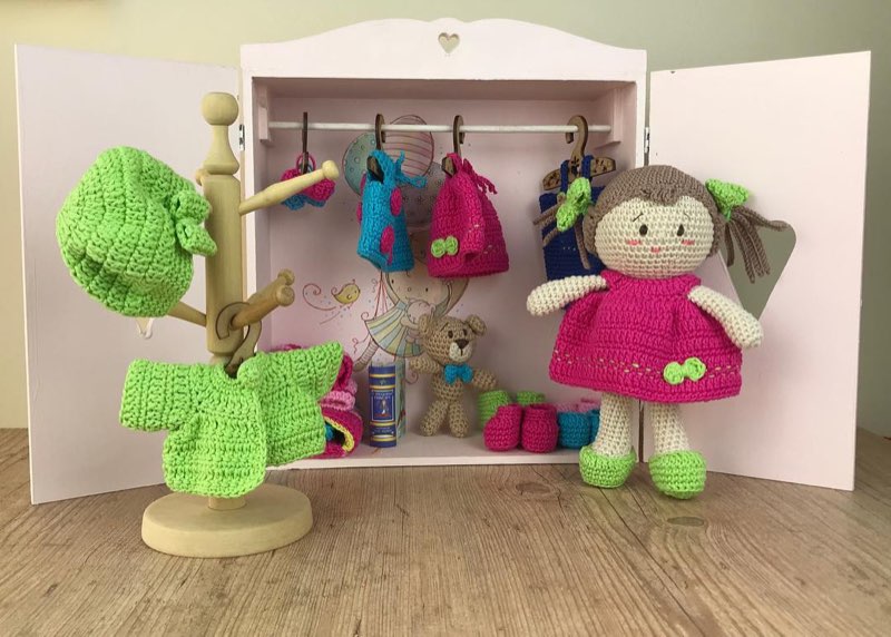 Boneca de crochê ao lado de um guarda-roupa em miniatura com roupinhas penduradas em cabides