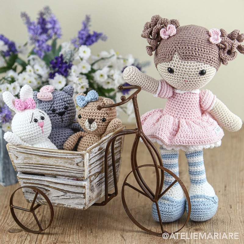 Boneca de crochê com vestido rosa ao lado de um triciclo em miniatura carregando uma coelha, um gatinho e um ursinho de amigurumi, com flores ao fundo