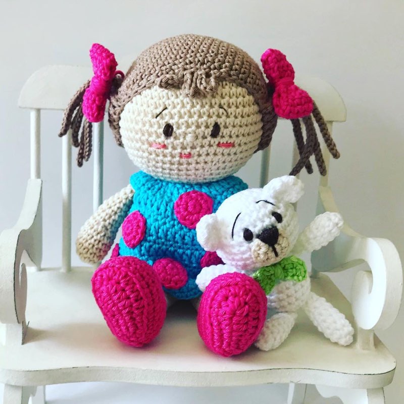 Boneca de crochê com vestido rosa-choque e detalhes de bolinhas, sentada ao lado de um cachorrinho branco de amigurumi