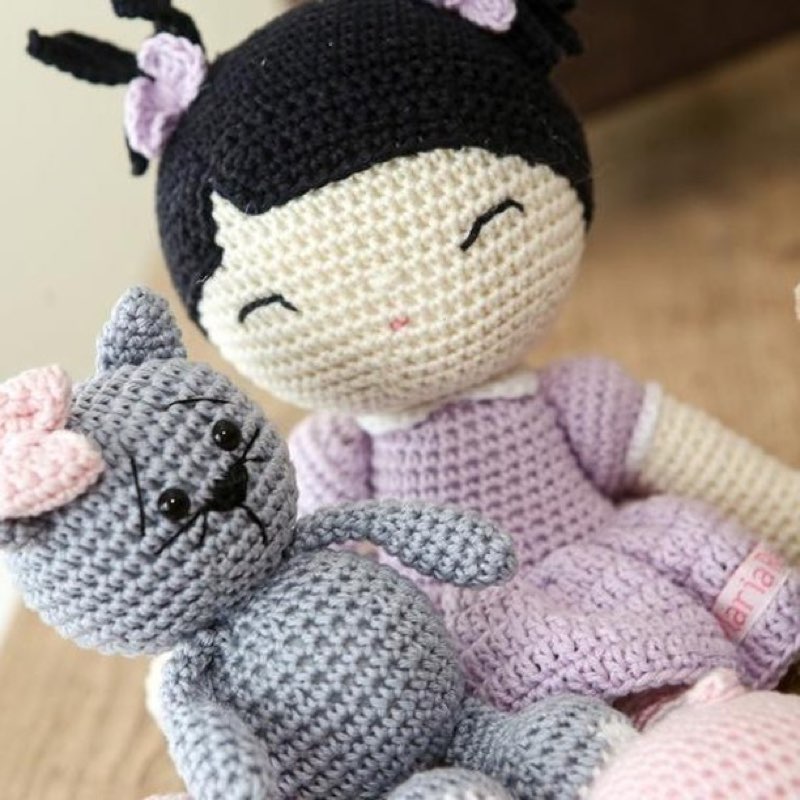 Boneca de crochê com vestido lilás e cabelo preto, sentada ao lado de um gatinho cinza de amigurumi