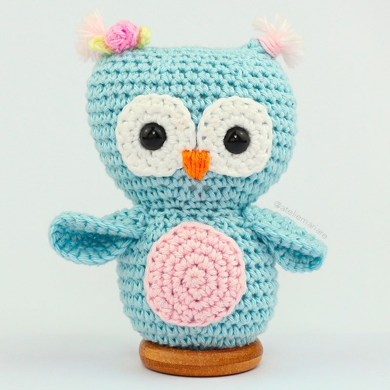Corujinha de crochê amigurumi nas cores azul-claro e rosa, em pé sobre uma base de madeira