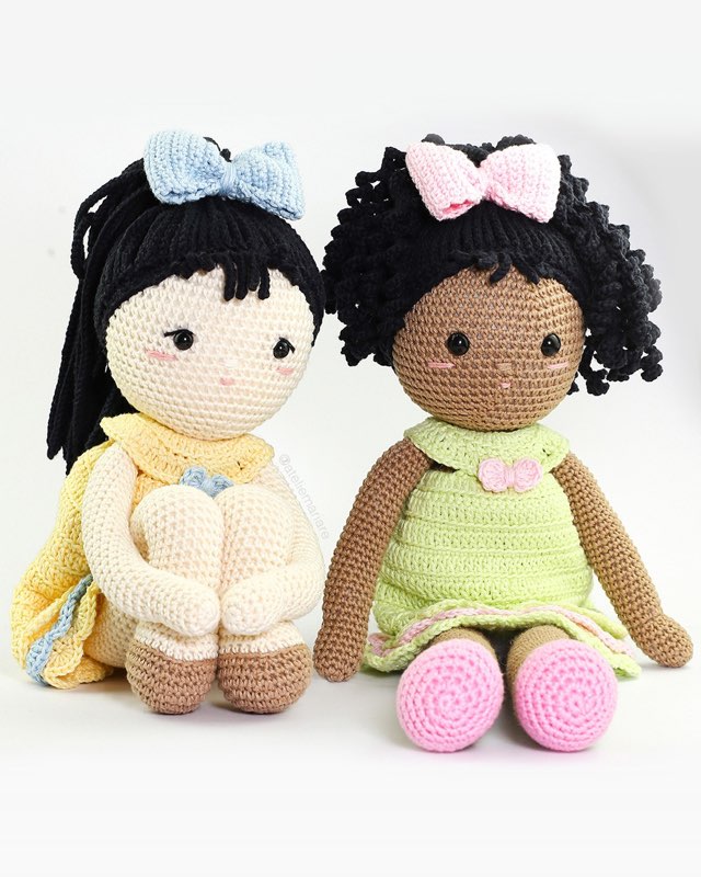 Duas bonecas de crochê de pele morena com vestidos amarelo e verde, sentadas juntas