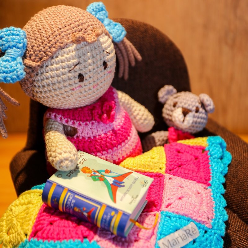 Boneca de crochê com roupinha rosa e ursinho de amigurumi, sentados sobre uma manta com livrinhos de crochê