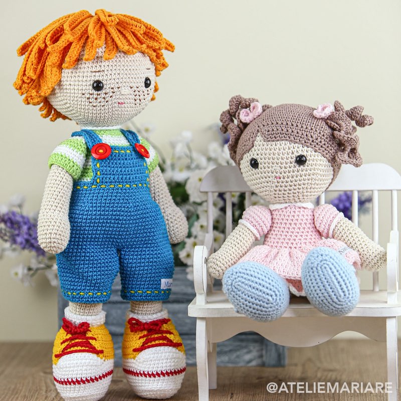 Boneco de crochê com cabelo cacheado laranja e jardineira azul, sentado ao lado de uma boneca com vestido rosa em um banquinho branco