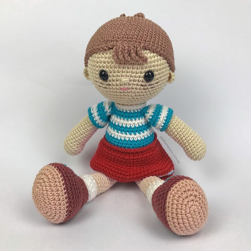 Boneco de crochê com camiseta listrada azul e branca e shorts vermelho, sentado sozinho