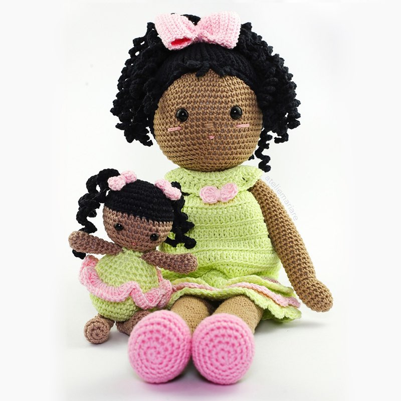 Boneca de crochê de pele morena com cabelo cacheado, vestido verde e laço rosa, sentada ao lado de sua mascotinha combinando