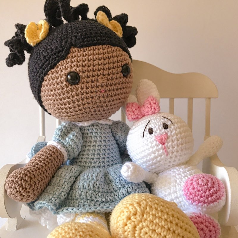 Boneca de crochê de pele morena com cabelo cacheado, sentada ao lado de um cachorrinho branco de amigurumi com laço azul
