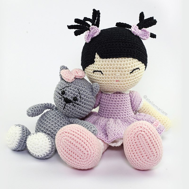 Boneca de crochê de pele morena com vestido lilás, sentada ao lado de um gatinho cinza de amigurumi