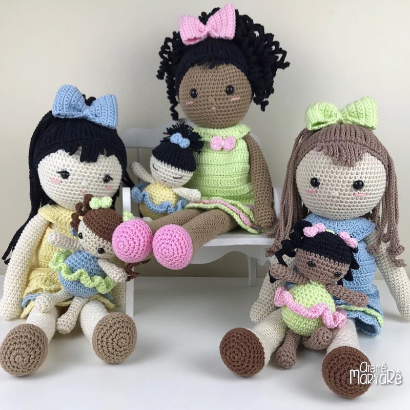 Três bonecas de crochê de pele morena com vestidos azul, verde e rosa, sentadas em um banquinho
