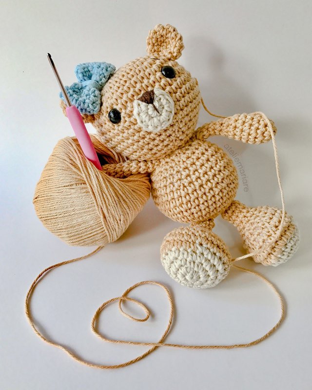 Boneco de crochê de um ursinho artesão segurando agulha de crochê ao lado de um novelo de lã